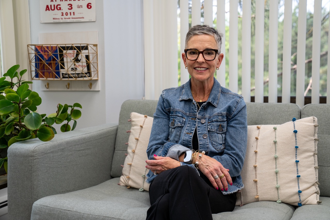 Stephanie Howey, a professional therapist with short gray hair and glasses, smiling warmly while sitting on a gray couch in a brightly lit counseling office at River's Bend, P.C.