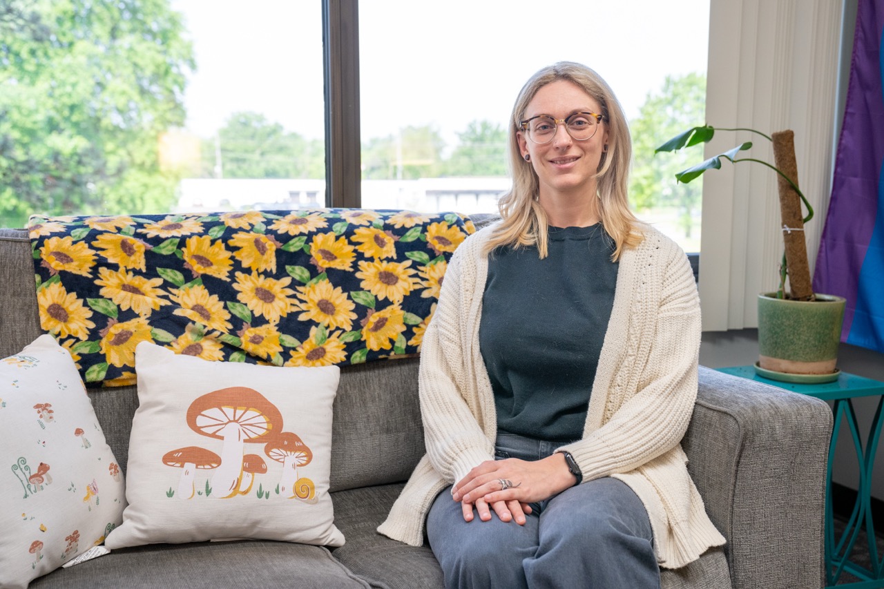 A smiling blonde female clinician Danielle Swantek, LMSW, CAADC wearing glasses and a cream cardigan sitting on a gray couch in a bright therapy room at River's Bend, P.C.