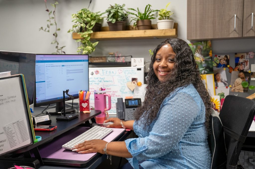 A smiling professional clinician at River's Bend, P.C. sitting at her office desk in front of dual monitors, ready to assist clients with substance abuse evaluations.