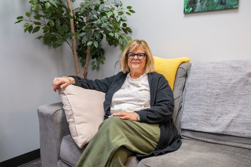 A smiling professional female therapist with short blonde hair and glasses sitting on a gray couch with a yellow accent pillow in a counseling office at River's Bend, P.C.