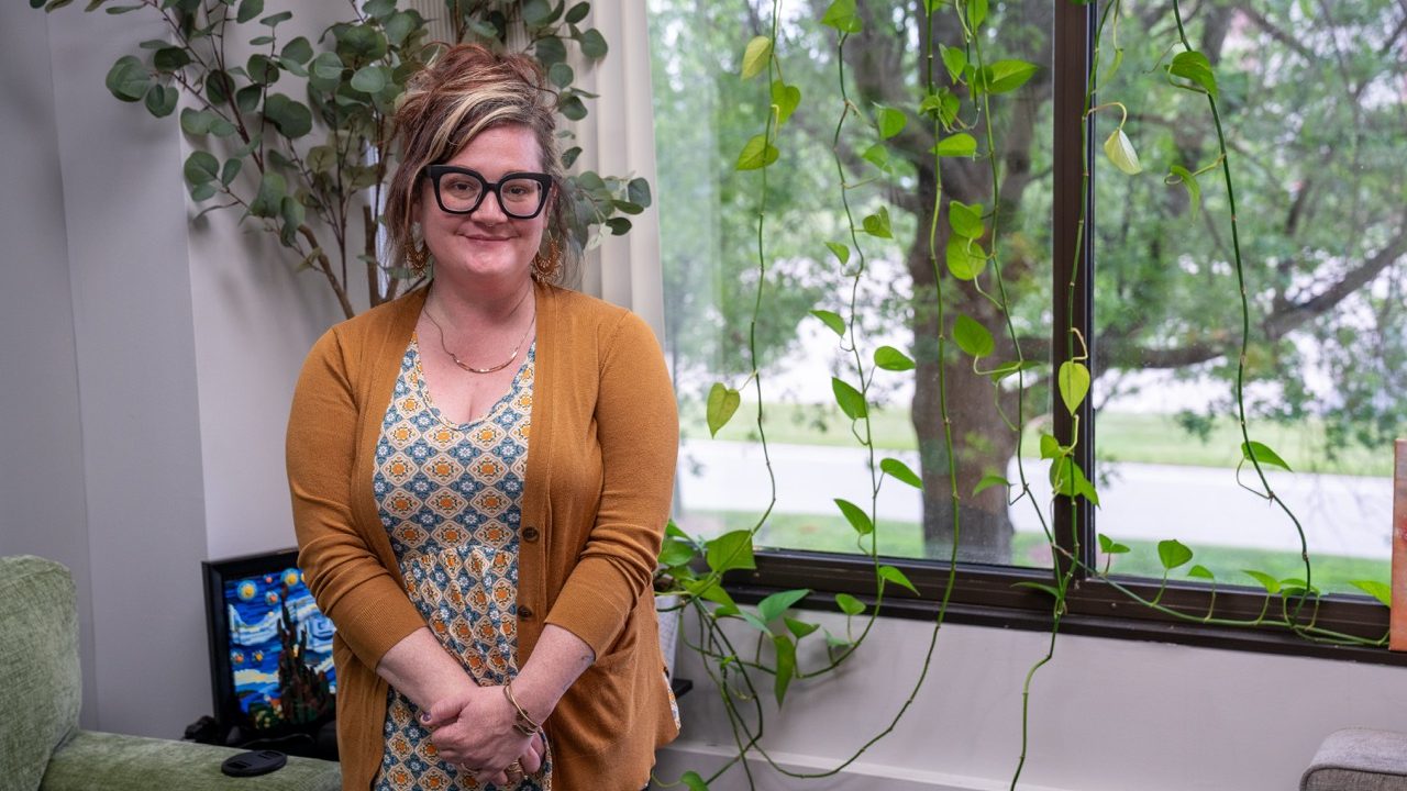 A professional and welcoming office desk setup featuring Katie, a clinician at River's Bend, P.C., smiling in front of a bright window with hanging pothos ivy plants.