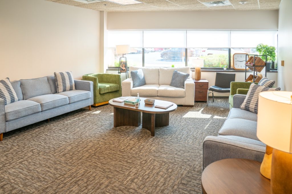 A welcoming, spacious group lounge area for PHP patients at River's Bend, P.C. with green and neutral couches and a large coffee table.