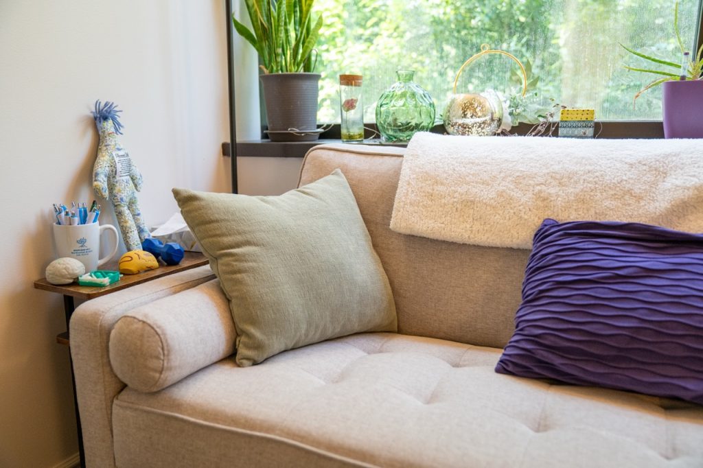 A comfortable, light gray tufted sofa in a brightly lit counseling office at River's Bend, P.C. with green plants and a large window in the background.