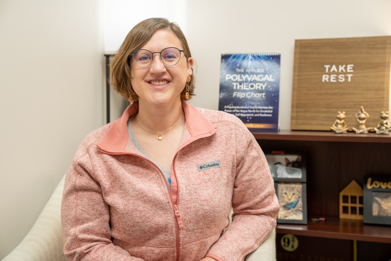 A smiling professional female clinician at River's Bend, P.C., wearing a pink fleece jacket, sitting in a counseling office next to a book titled "The Applied Polyvagal Theory Flip Chart."