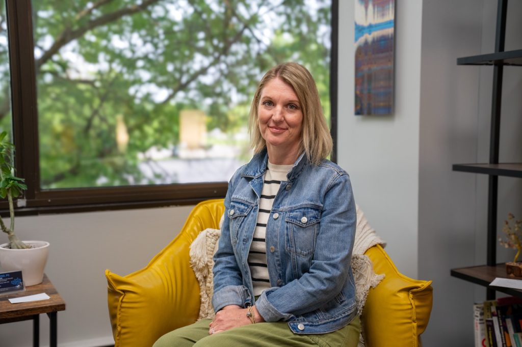 A smiling female therapist Amy Fresch sitting in a yellow armchair inside a brightly lit office at River's Bend, P.C.