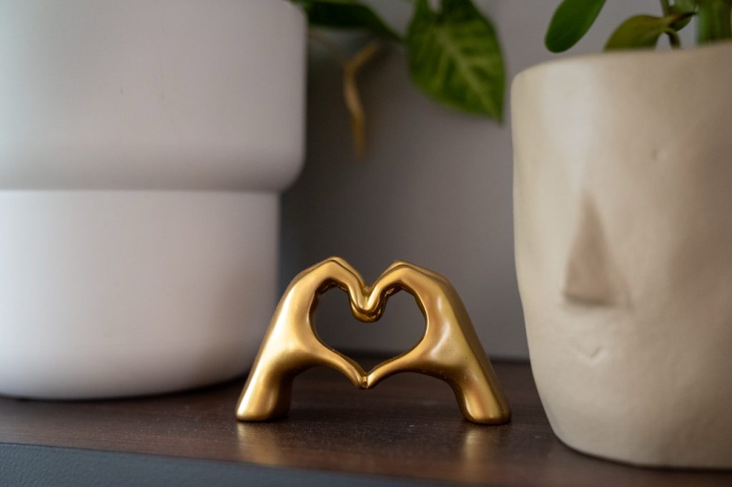 A gold decorative sculpture forming a heart shape with hands, sitting on a wooden shelf next to a modern face-shaped ceramic planter in a counseling office at River's Bend, P.C.