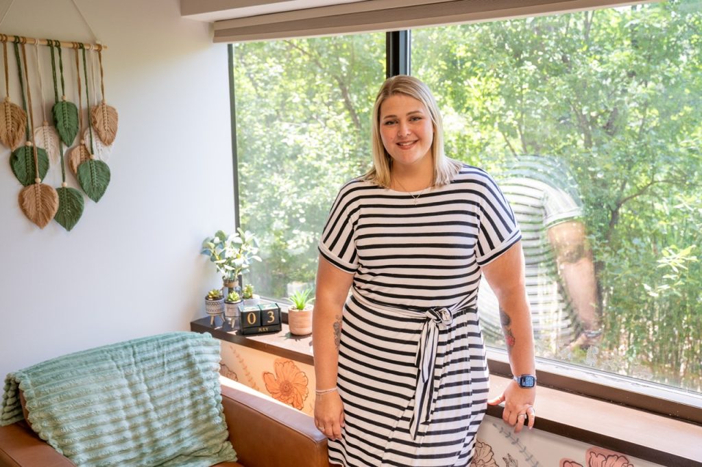 A smiling Jessica Hillen Clinical Director, wearing a striped dress standing in a bright therapy room at River's Bend, P.C.
