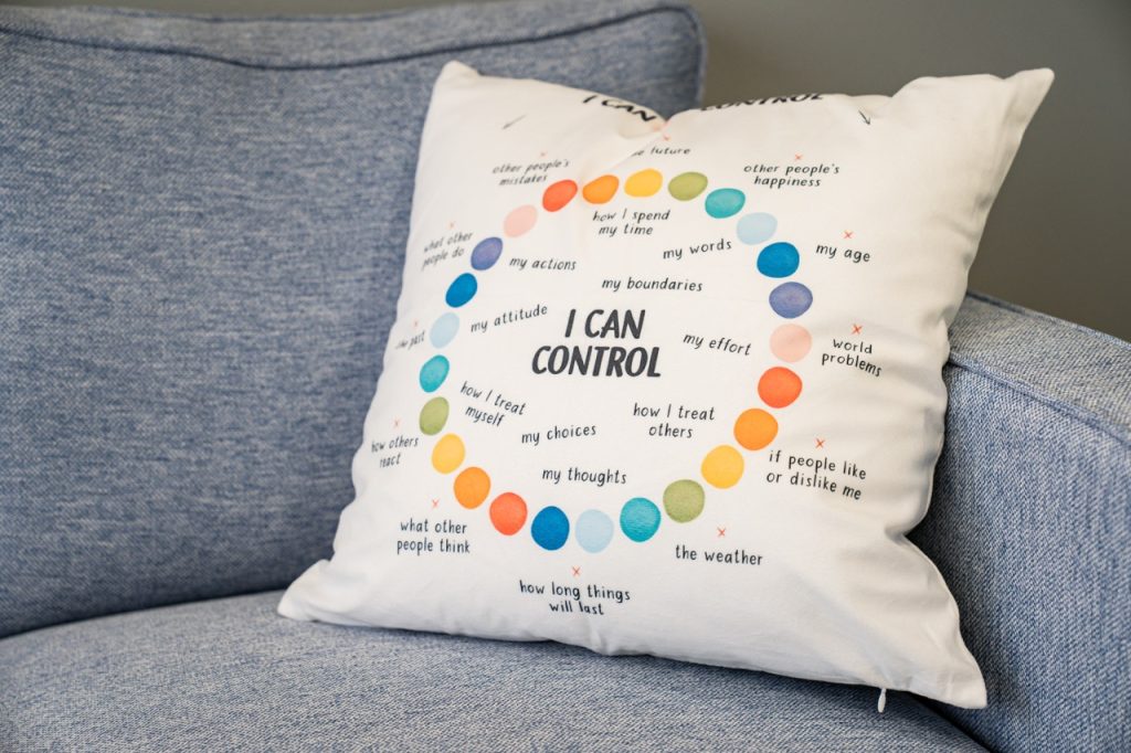 A close-up of a decorative throw pillow on a blue office sofa, featuring an educational graphic titled "I Can Control" surrounded by colorful circles highlighting personal choices and mindset.