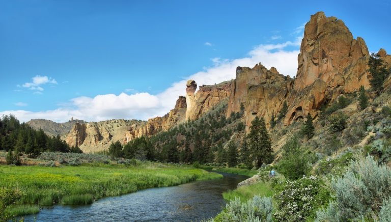 A wide, scenic view of a winding green river flowing through the rocky canyon and towering cliffs of Smith Rock Park under a clear sky.