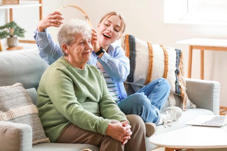 A young woman smiles warmly as she puts headphones on her grandmother at home, capturing a tender moment of connection between generations.