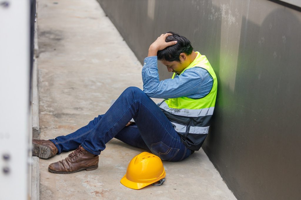 A tired construction worker wearing a safety vest and helmet sits on the ground at a job site, resting his head in his hand in distress.