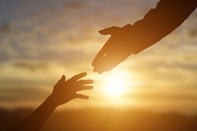 Two hands reaching toward each other to hold hands, back-lit by warm golden sunlight during sunset.