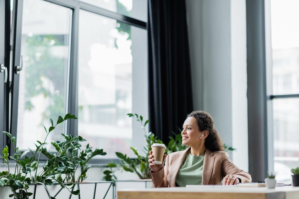 Woman sitting at a office desk holding a coffee cup and looking out the window while taking a break from her computer.