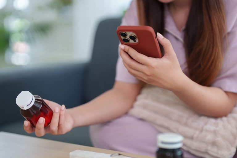 Woman holding a smartphone and a prescription medicine bottle while looking up healthcare information online.
